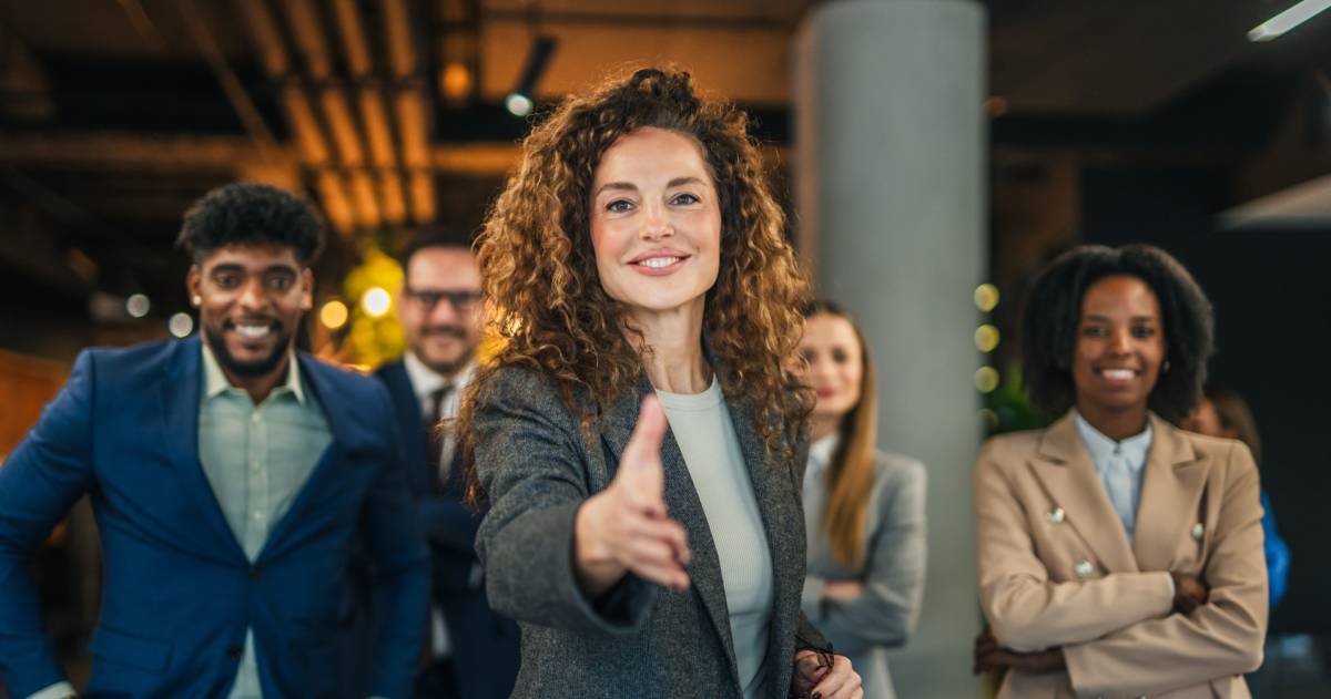 Een zelfverzekerde zakenvrouw biedt een hand aan, met een divers team van zakenmensen achter haar.