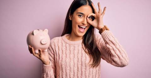 Vrolijke jonge vrouw met een spaarvarken in haar hand en met haar andere hand maakt ze een ok teken voor haar oog staat voor een roze achtergrond